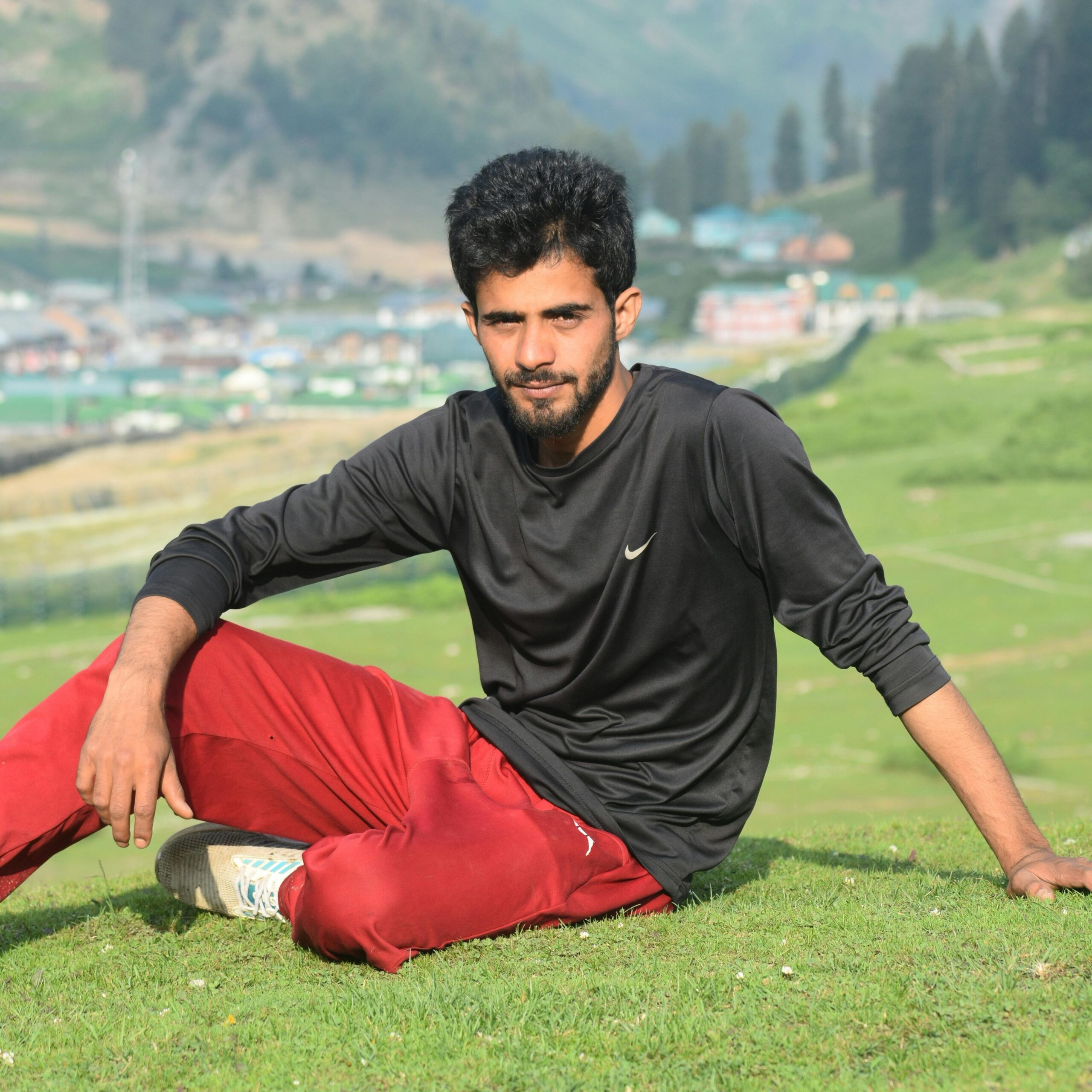 A young man in casual attire sits on a grassy hill with a beautiful mountainous backdrop.