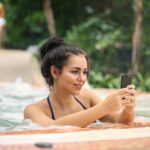 Smiling woman in a bikini enjoying a jacuzzi while texting on her smartphone outdoors.